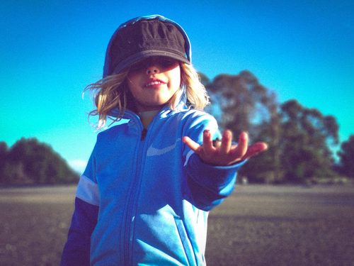 Mid-shot portrait of a child taken from a low angle, wearing a hat that obscures their eyes, with an open palm reaching toward the camera against a blue sky.